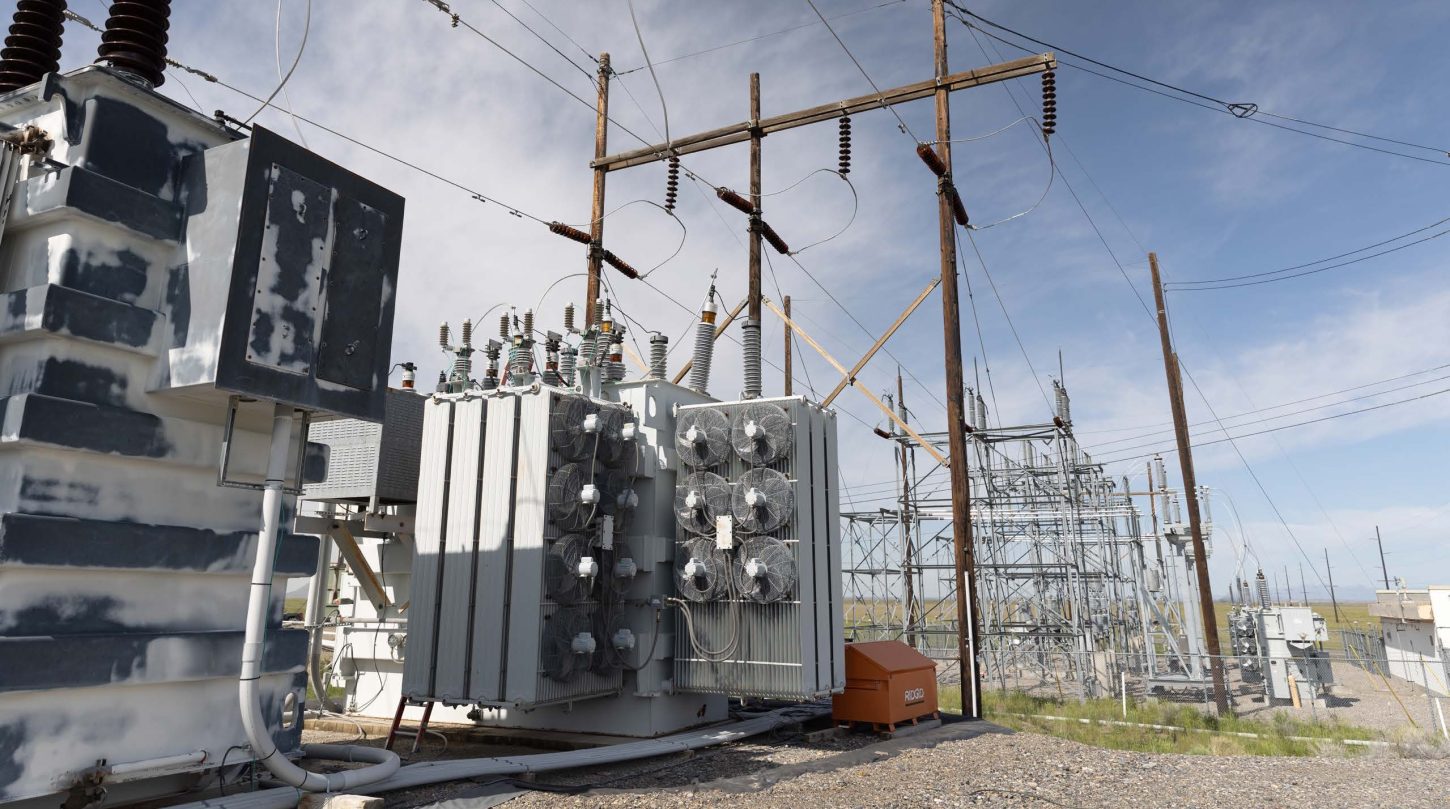 Electrical transformers and cooling fans at an outdoor power substation with utility poles and transmission lines.