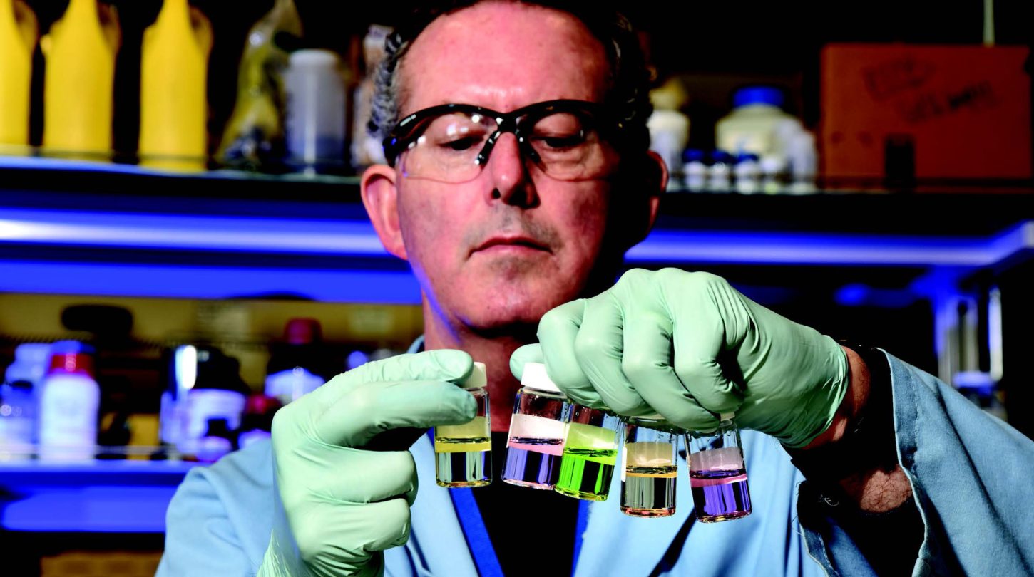 Scientist wearing safety glasses and gloves examines several small vials containing colored liquid samples in a laboratory.