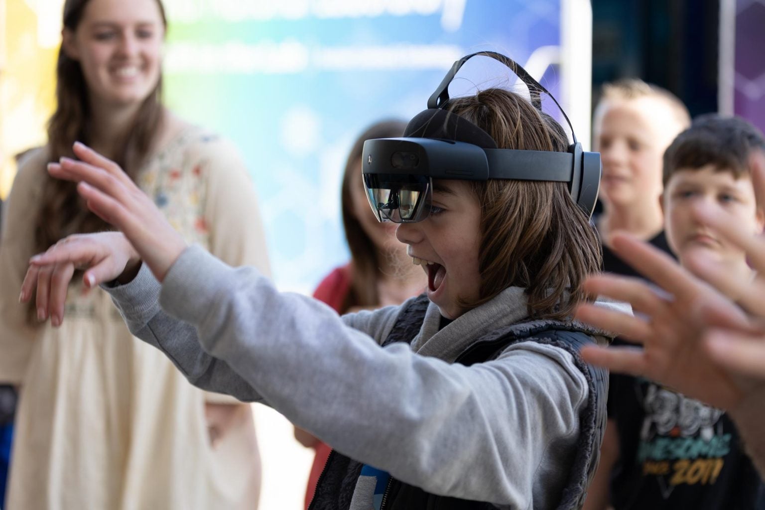 Child wearing a VR headset, reaching out excitedly while classmates and a teacher look on.