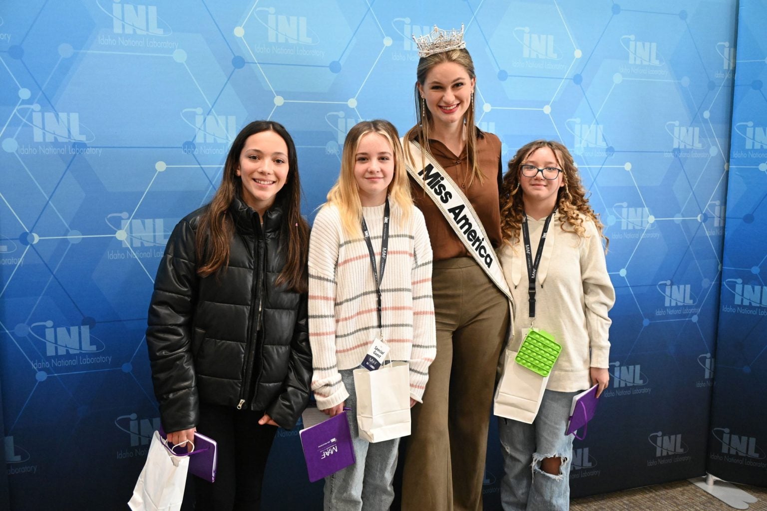 Miss America posing with three girls in front of an Idaho National Laboratory backdrop.