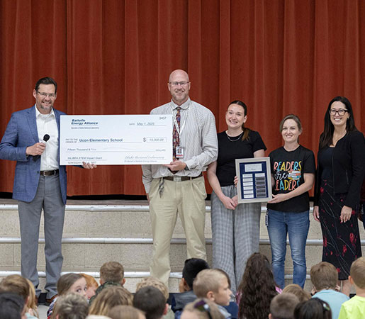 Group presenting a large donation check to a school in front of an audience of students.
