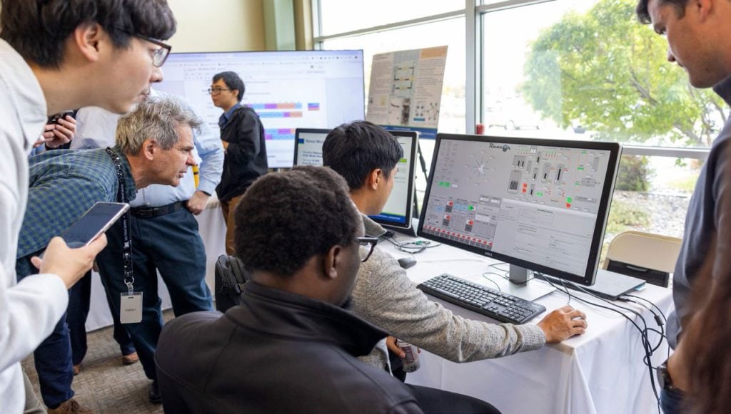 Attendees gather around computer workstations to observe a demonstration of a digital control system during a technical research event.