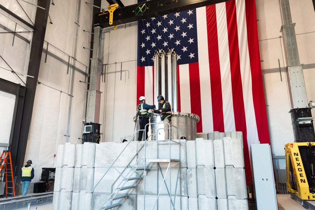 Two workers at Aalo Atomics' wearing safety harnesses stand on a platform installing equipment inside a high-bay facility, with a large U.S. flag hanging behind the assembly.