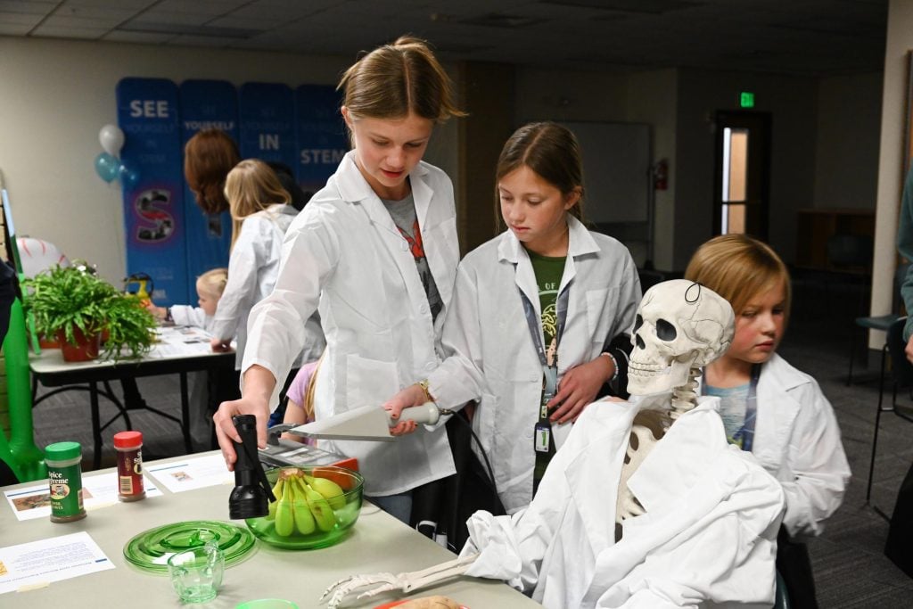 Students in lab coats participate in a hands-on STEM activity with a skeleton model and other science materials.