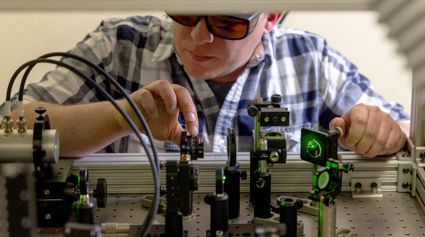 Researcher wearing safety glasses adjusts optics on a green laser setup.