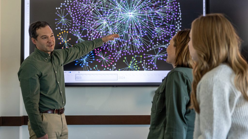 INL researcher points to a network data visualization on a large screen as two colleagues observe.